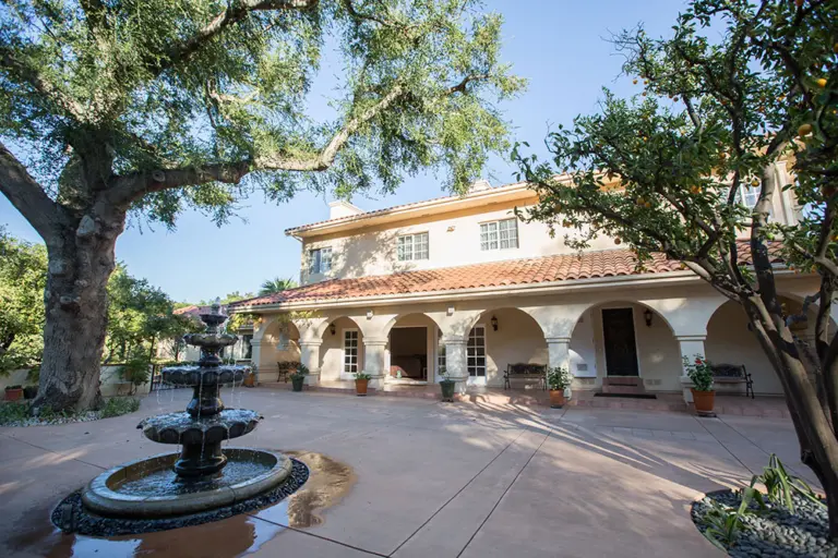 Two-story cream stucco building with arched breezeway, courtyard fountain, and large oak tree