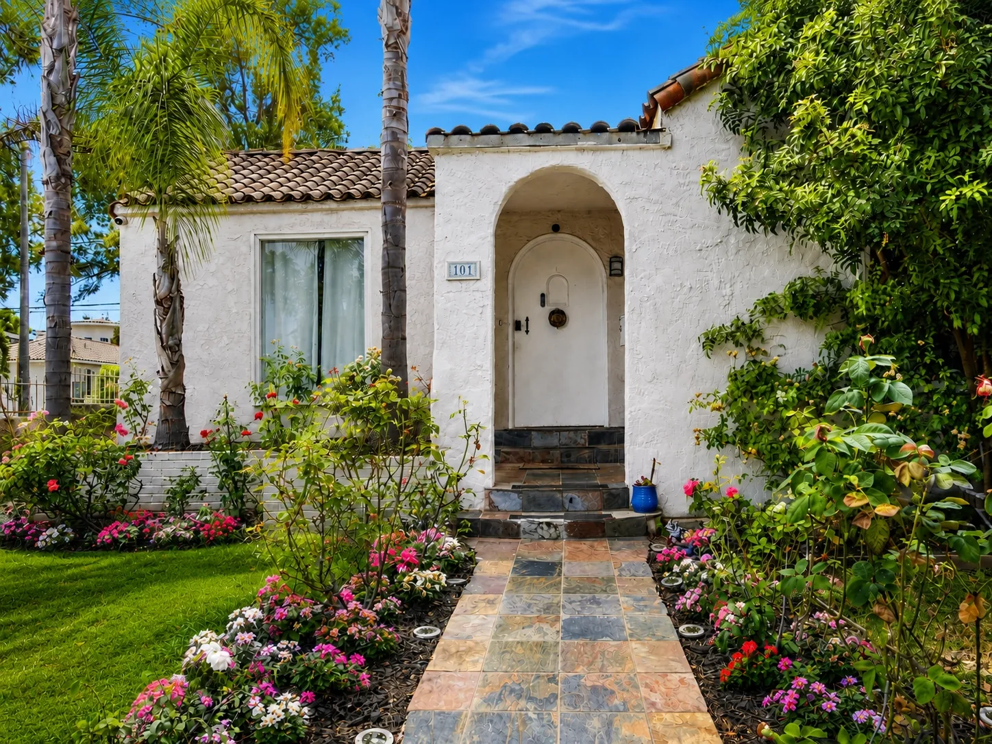White stucco house with an arched entryway, tiled walkway, and flower beds under a blue sky