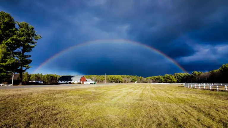 Open pasture with a red barn, white fencing and a rainbow over a wooded horizon