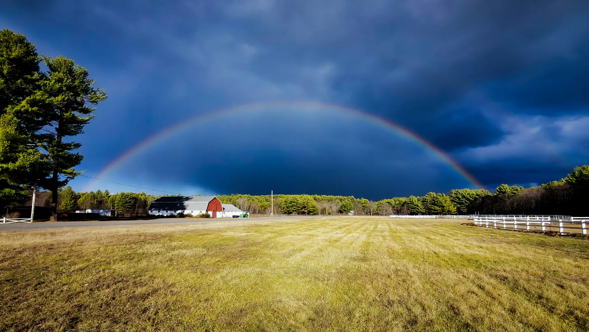 Open pasture with a red barn, white fencing and a rainbow over a wooded horizon