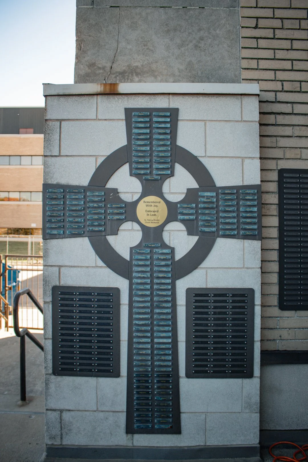 St. Patrick Center client memorial cross mounted on a building wall, with a plaque reading Remembered With Joy