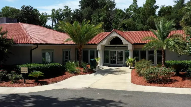 Single-story tile-roofed building with a Saint Simons By-The-Sea entrance sign and a palm-lined walkway