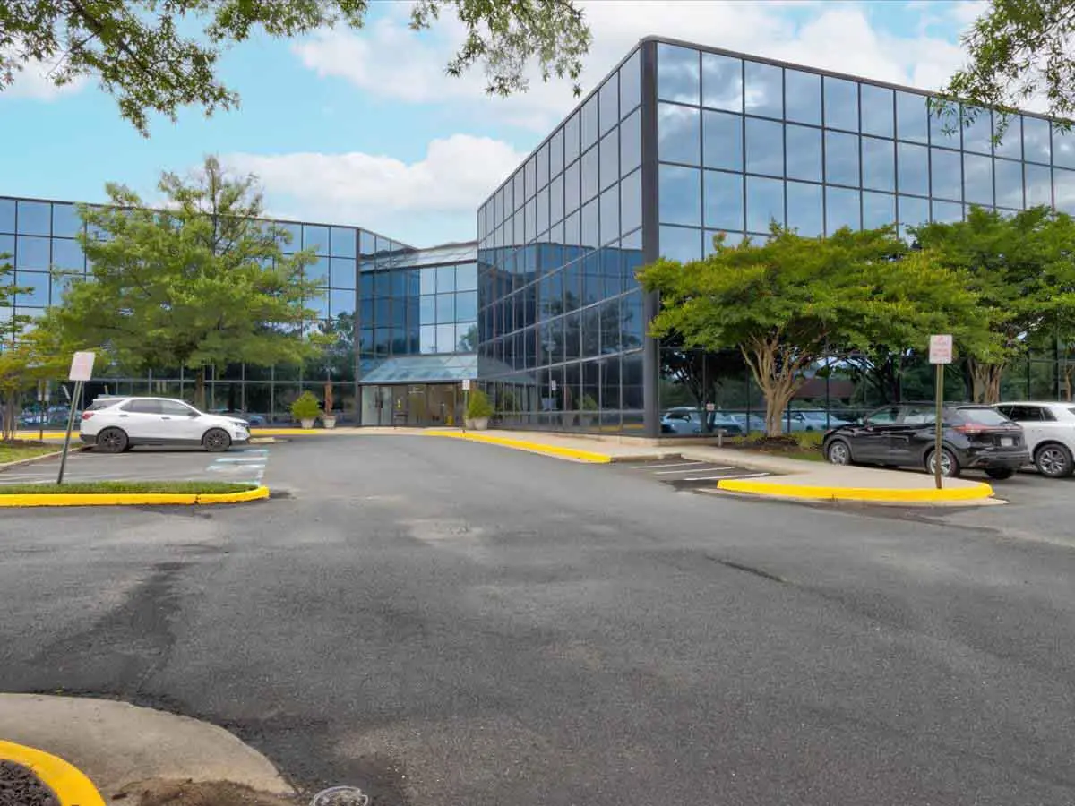 Glass-walled office building with a covered entrance and a parking lot in front
