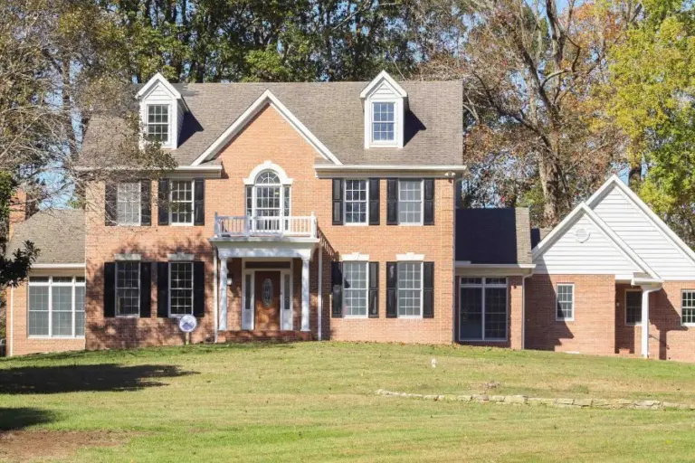 Two-story brick colonial house with black shutters and white trim, set on a lawn with trees behind it