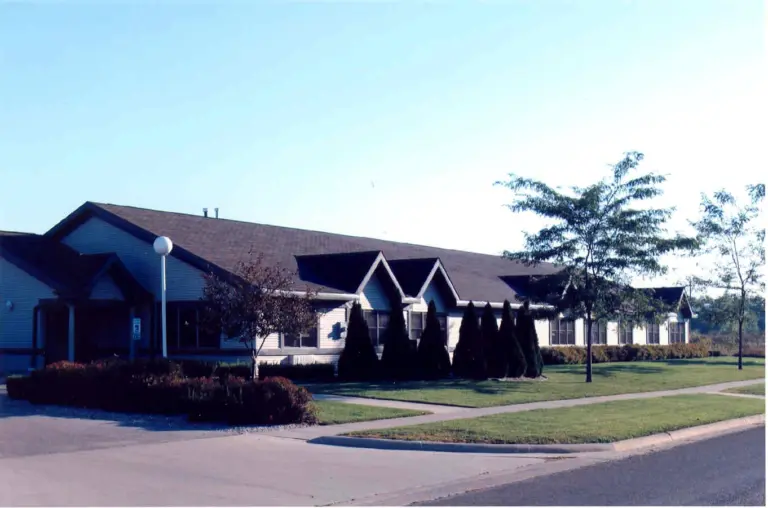 Single-story building with light siding, shrubs, and trees along a paved driveway