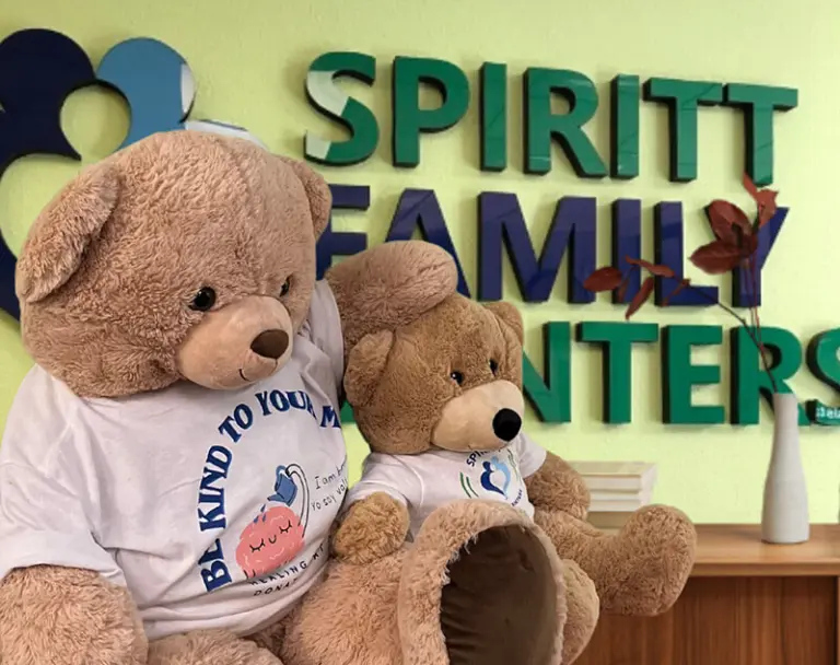 Two plush teddy bears in a lobby in front of a green and blue SPIRITT Family Centers wall sign