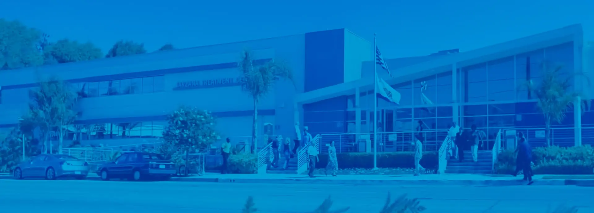 Blue-tinted two-story building with entrance steps, a flagpole, and people walking outside