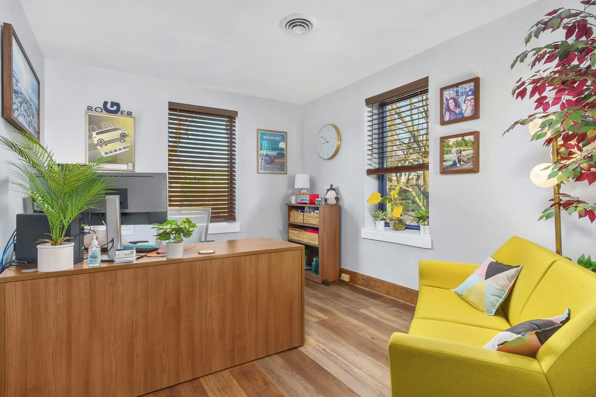 Furnished office with a wood desk, yellow sofa, plants and framed pictures near a window