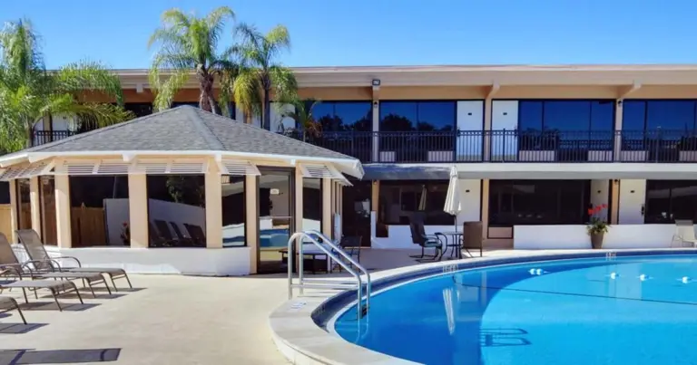 Two-story beige building with a pool and gazebo-style structure under palm trees
