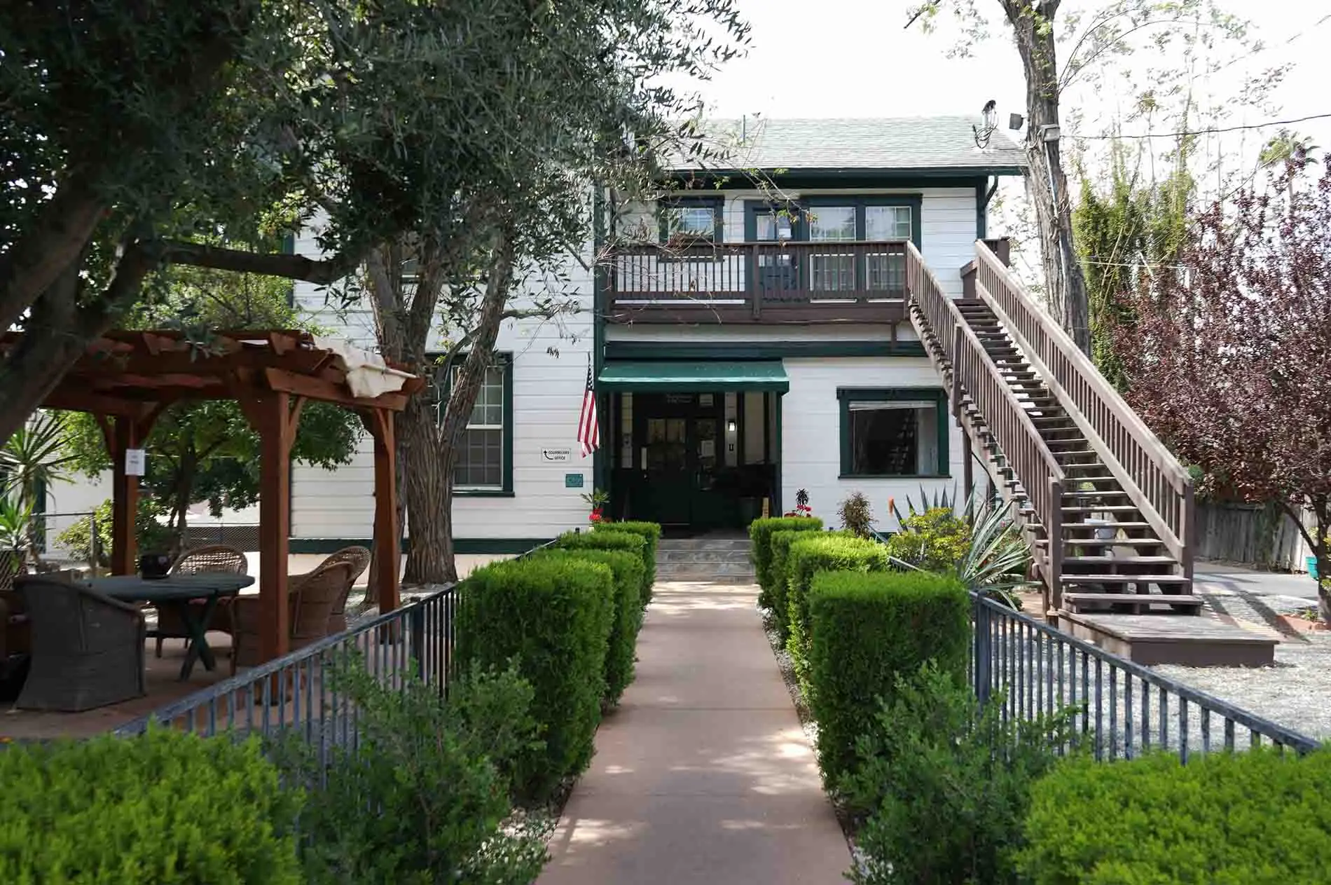 Two-story white building with green awning, staircase, and hedged walkway